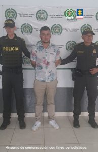 En la imagen se ve una persona detenida bajo custodia de dos integrantes de la Policía Nacional. Detrás suyo un backing institucional.