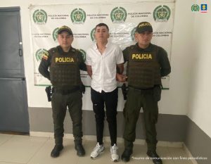 en la imagen se ve una persona detenida bajo custodia de dos integrantes de la Policía Nacional. Detrás suyo un backing institucional.