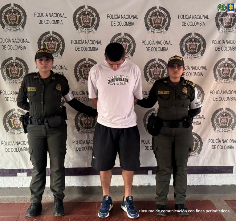 en la imagen se ve un hombre detenido bajo custodia de dos uniformados de la Policía Nacional. Detrás suyo, un backing institucional.