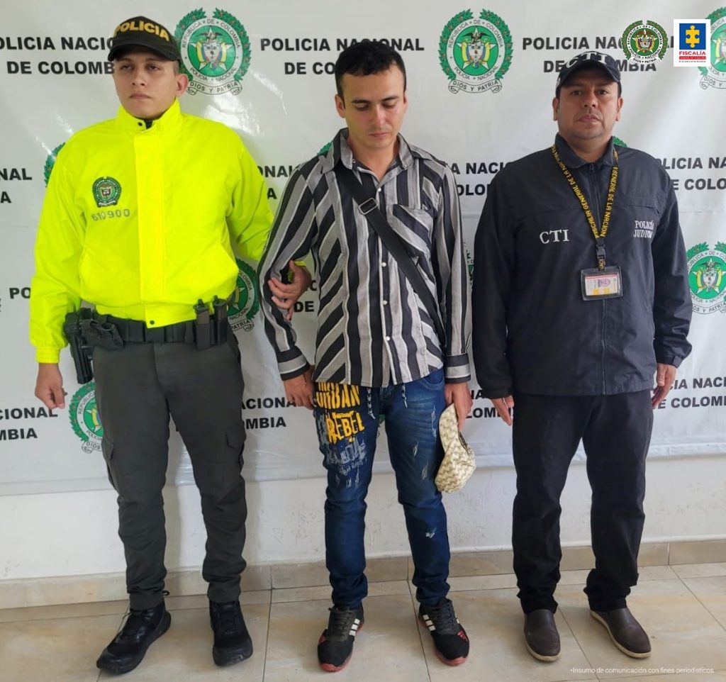 En la imagen se ve una persona detenida en custodia de un integrante de la Policía Nacional y del CTI. Detrás suyo un backing de la Policía.