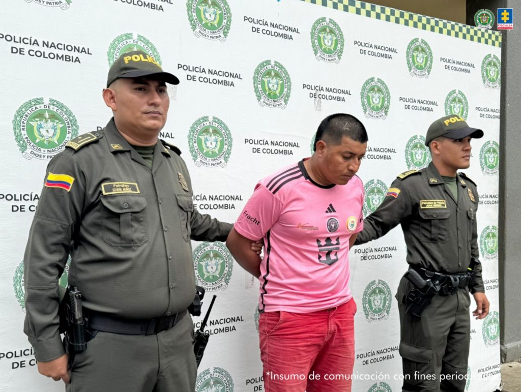en la imagen se ve una persona detenida bajo custodia de dos uniformados de la Policía Nacional. Detrás de ellos un backing oficial.