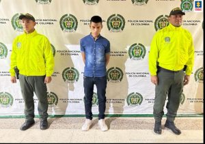 en la imagen se ve una persona detenida bajo custodia de dos integrantes de la Policía Nacional. Detrás suyo un backing institucional.