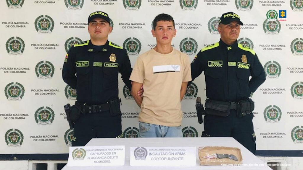en la fotografia se puede observar al capturado en medios de dos policias, en frente de ellos hayun mesa con los elementos incautados y detras un baking de la policia
