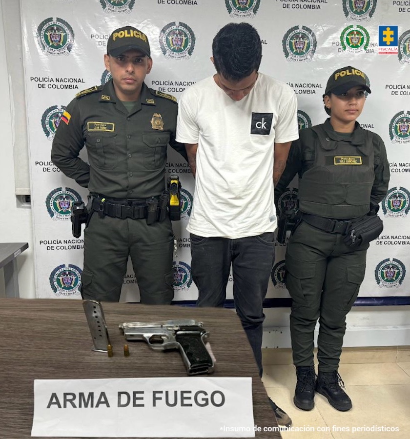 en la imagen se ve un hombre detenido bajo custodia de dos integrantes de la Policía Nacional. Delante suyo una mesa con un arma de fuego. Detrás un backing institucional.