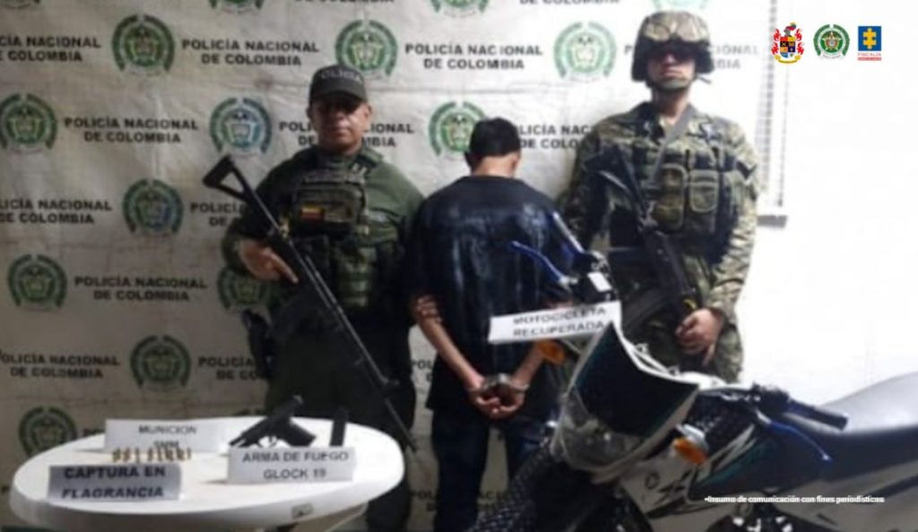 en la imagen se ve un hombre detenido bajo custodia de dos integrantes de la Policía Nacional. Delante de él una mesa con elementos incautados y a su lado, una motocicleta. Detrás, un backing institucional.