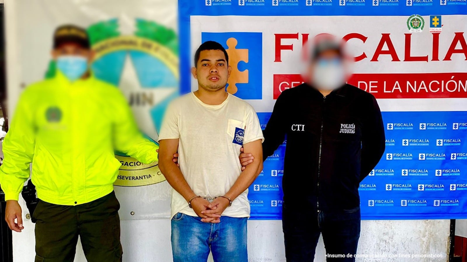 Se observa al capturado junto a un uniformado de la Policía Nacional e investigador del CTI. Detrás los banner que identifican a la Policía Nacional y a la Fiscalía General de la Nación.