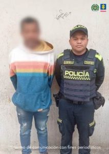 En la imagen se observa a dos personas de pie frente a una pared de color claro.A la derecha está un hombre vestido con uniforme de la Policía Nacional de Colombia, que incluye chaleco antibalas negro con la palabra “POLICÍA” en letras amarillas. También lleva gorra del mismo cuerpo policial. A la izquierda hay otra persona, con el rostro difuminado, que viste un buso con franjas horizontales de colores (naranja, blanco, azul y verde) y pantalón de mezclilla roto en las rodillas. En la parte superior derecha de la imagen aparecen los logotipos de la Policía Nacional de Colombia y la Fiscalía General de la Nación. En la parte inferior, en letras pequeñas, se lee: “Insumo de comunicación con fines periodísticos”.