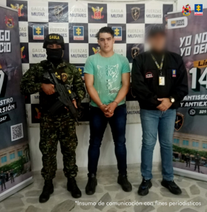 Se visualiza al capturado junto a un uniformado del Ejército Nacional y un investigador del CTI. Detrás de ellos los banners que identifican al Ejército Nacional.