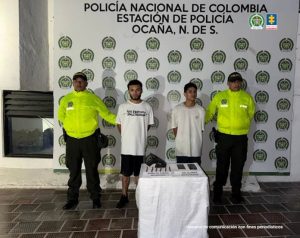 Se visualiza a los capturados junto dos uniformados de la Policía Nacional. Frente a ellos una mesa con el arma, la munición y los teléfonos incautados. Detrás de ellos el banner que identifica a la Policía Nacional.