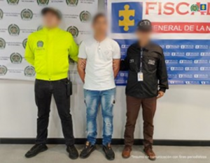 Se visualiza al capturado junto a un uniformado de la Policía Nacional y un investigador del CTI con los rostros difuminados. Detrás de ellos los banners institucionales que identifican a la Policía Nacional y a la Fiscalía General de la Nación.