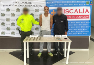 Se visualiza al capturado junto a un uniformado de la Policía Nacional y un investigador del CTI. Frente a ellos una mesa con las sustancias incautadas. Detrás de ellos los banners que identifican a la Policía Nacional y a la Fiscalía General de la Nación.