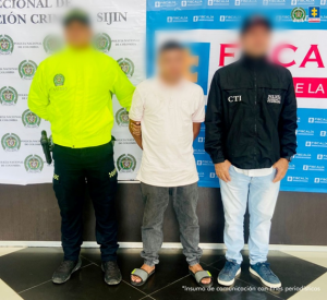Se visualiza al capturado junto a un uniformado de la Policía Nacional y un investigador del CTI. Detrás de ellos se ubica los banners que identifican a la Policía Nacional y a la Fiscalía