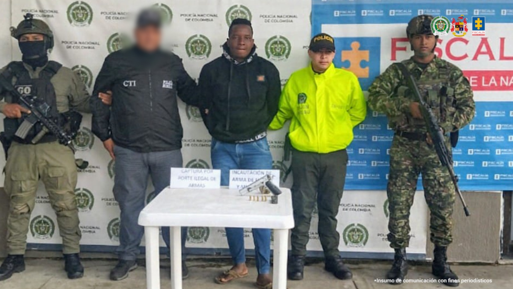 En la fotografía aparece el capturado junto a un funcionario del CTI, dos uniformados de la Policía y un soldado del Ejército. En la parte superior están un banner de la Fiscalía General y otro banner de la Policía