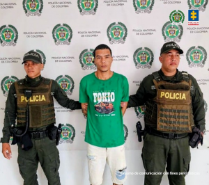 En la imagen se observa a un hombre en el centro, vestido con una camiseta verde que tiene un estampado que dice “TOKIO” y un pantalón claro, siendo custodiado por dos policías uniformados de la Policía Nacional de Colombia.Ambos agentes llevan chalecos antibalas con la palabra POLICÍA en color amarillo y están de pie a los lados del detenido, sujetándolo por los brazos. El fondo es un mural institucional con logotipos de la Policía Nacional de Colombia y, en la esquina superior derecha, aparecen los logotipos de la Policía Nacional y la Fiscalía General de la Nación. En la parte inferior derecha se lee un texto pequeño: “Insumo de comunicación con fines periodísticos”, lo que indica que la foto es de carácter oficial y utilizada para comunicación pública.