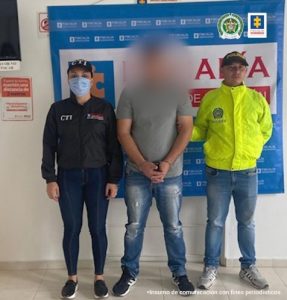 En la imagen se observa al capturado con el rostro difuminado junto a una investigadora del CTI y a un integrante de la Sijin de la Policía Nacional. Detrás de ellos el banner que identifica a la Fiscalía General de la Nación.