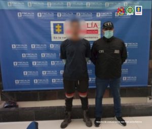 En la imagen se observa el capturado con el rostro difuminado junto a un investigador del CTI. En la parte posterior está ubicado el banner que identifica a la Fiscalía General de la Nación.