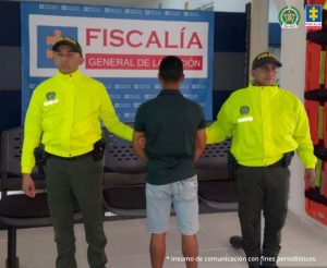 Se observa al capturado de espaldas junto a dos uniformados de la Policía Nacional. Detrás el banner institucional de la Fiscalía General de la Nación