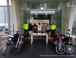 Se visualiza a cinco de los capturados junto a dos uniformados de la Policía Nacional. Frente a ellos los elementos incautados (arma – motocicletas). Detrás se ubican el banner que identifica a la Policía Nacional.