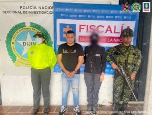 En la imagen se aprecia al capturado junto a una uniformada de la Policía Nacional, una investigadora del CTI y a un uniformado del Ejército Nacional. Detrás de ellos se observa los banners que identifican a la Policía Nacional y a la Fiscalía General de la Nación.