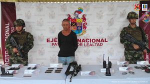 En la fotografía aparece el capturado junto a uniformados del Ejército Nacional. Frente a ellos se ubica una mesa con el material incautado: armas, municiones, proveedores, radios de comunicación y teléfonos celulares.