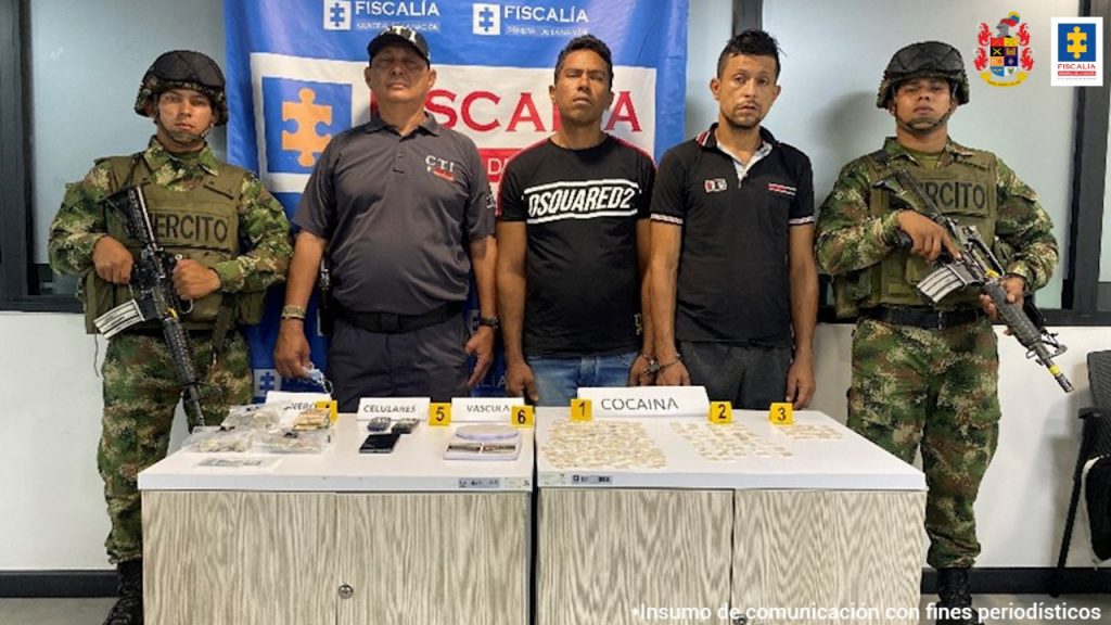 En la fotografía se observa a los dos capturados junto a un investigador del CTI y dos uniformados del Ejército Nacional, frente a ellos una mesa con los elementos incautados. En la parte posterior el banner que identifica a la Fiscalía General de la Nación.