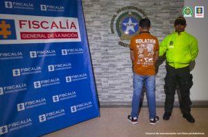 En la imagen se observa al capturado de espaldas junto a un uniformado de la Policía Nacional. Detrás de ellos se visualizan los banners de la Fiscalía General de la Nación y la Policía Nacional.