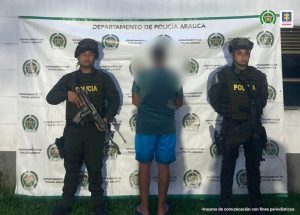 En la fotografía se visualiza el capturado de espaldas junto a dos uniformados de la Policía Nacional. En la parte posterior se ubica el banner que identifica a la Policía Nacional.