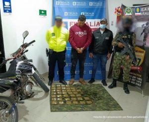 Se visualiza al capturado junto a un investigador del CTI uniformados de la Policía y Ejército Nacional. Al frente en el piso se ubicó la munición incautada. Detrás de ellos el banner que identifica a la Fiscalía General de la Nación y al Ejército Nacional.