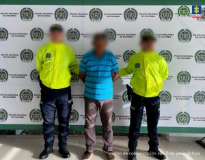 Se visualiza al capturado junto a dos uniformados de la Policía Nacional con el rostro difuminado. Detrás se ubica el banner que identifica a la Policía Nacional.