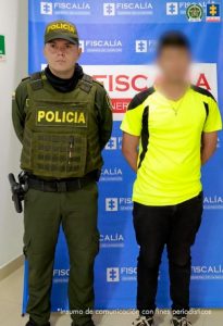 Se visualiza al capturado con el rostro difuminado junto a uniformado de la Policía Nacional. Detrás se ubica el banner de la Fiscalía General de la Nación.