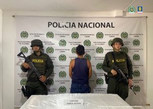 Se observa al capturado de espalda acompañado de dos uniformados de la Policía Nacional. Detrás el banner institucional que identifica a la Policía Nacional