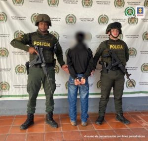 En la imagen se observa al investigado de espaldas junto a dos uniformados de la Policía Nacional. Detrás de ellos el banner que identifica al Departamento de Policía Arauca