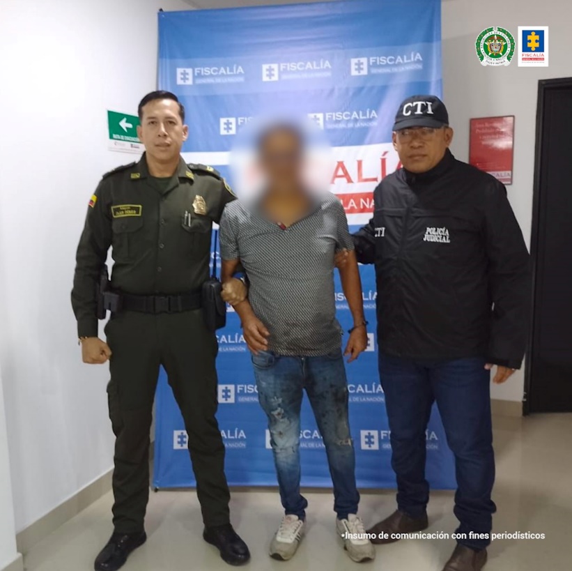 En la imagen se aprecia el capturado junto a un investigador del CTI y un uniformado de la Policía Nacional.  En la parte posterior se observa el banner que identifica La Fiscalía General de la Nación.