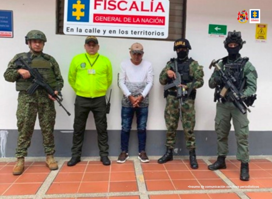 En la imagen se observa al capturado junto a uniformados del Ejército y de la Policía Nacional. Detrás de ellos un banner que identifica a la Fiscalía General de la Nación