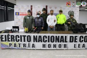 En la imagen se observa al capturado junto a un investigador del CTI y uniformados de la Fuerza Aérea, Ejército y Policía Nacional. Frente a ellos una mesa con los elementos incautados y un banner que identifica al Ejército Nacional. Se observa de tras de ellos banners que identifican a la Fiscalía General de la Nación, Ejército y Policía Nacional