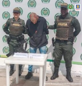 En la imagen se observa el hombre junto a dos uniformados de la Policía Nacional. Frente a ellos una mesa donde se ubico el arma y munición incautada. En la parte posterior se encuentra el banner que identifica a la Policía Nacional.