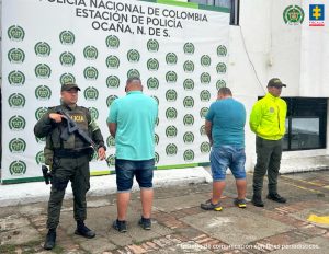 Se observa a los dos capturados de espaldas junto a dos uniformados de la Policía Nacional. Detrás de ellos el banner que identifica a la Policía Nacional.