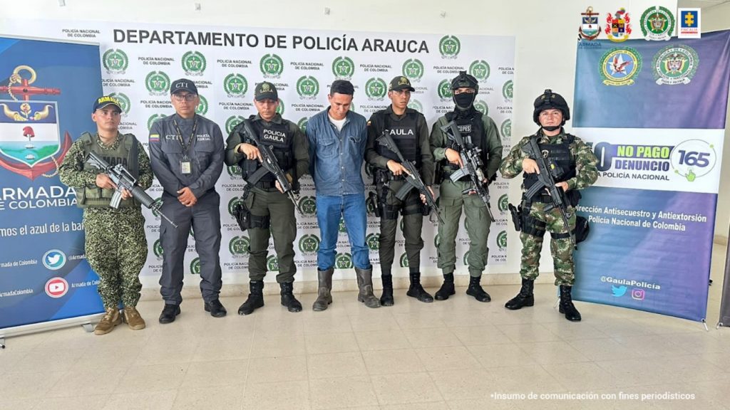 En la imagen se aprecia el capturado junto a un investigadore del CTI y uniformados de la Policía, Ejército y Armada Nacional. En la parte posterior se observan los banners de la Policía y Armada Nacional.