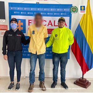 En la imagen se aprecia al capturado junto a una investigadora del CTI y a un integrante de la Sijin. En la parte posterior se observa el banner que identifica a la Fiscalía General de la Nación y la bandera de Colombia.