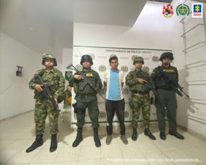 En la imagen se observa al capturado junto a uniformados de la Policía y Ejército Nacional. En la parte posterior se observa el banner que identifica al Departamento de la Policía Nacional