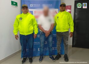 Se observa al capturado con el rostro difuminado junto a dos uniformados de la Policía Nacional. En la parte posterior se encuentra el banner que identifica a la Fiscalía General de la Nación.