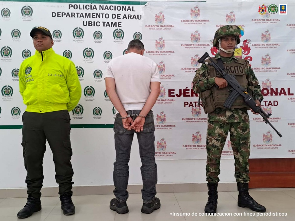 En la imagen se observa al capturado de espaldas junto a dos uniformados de la Policía y Ejército Nacional. Detrás de ellos los banners que identifican a la Departamento de Policía de Tame y al Ejército Nacional.