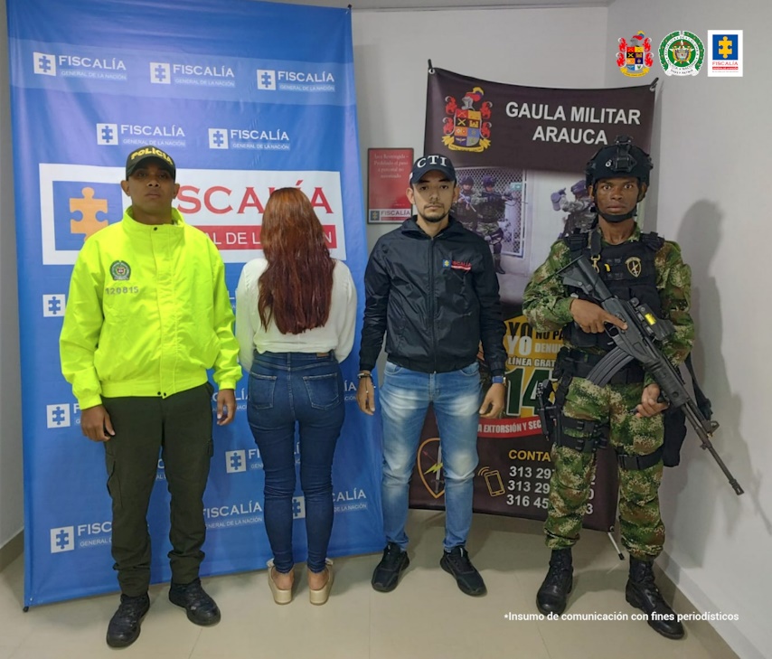 En la imagen se observa la capturada de espaldas junto a un investigador del CTI y uniformados de la Policía y Ejército Nacional. En la parte posterior se observan los banners que identifican a la Fiscalía General de la Nación y al Gaula Militar.