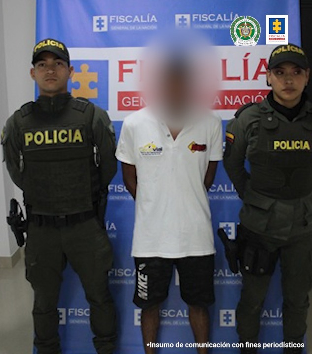 En la imagen se observa al capturado con el rostro difuminado junto a dos uniformados de la Policía Nacional. Detrás de ellos el banner que identifica a la Fiscalía General de la Nación.