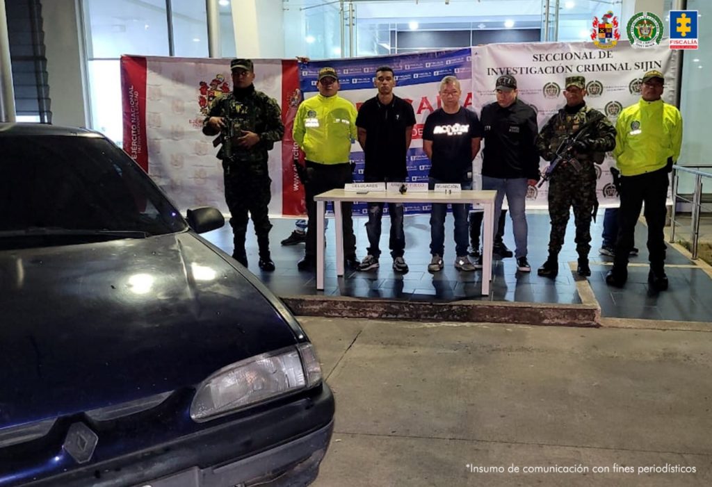 Se visualiza a los capturados junto a un investigador del CTI y uniformados de Ejército y Policía Nacional. Frente a ellos en una mesa los elementos incautados. Detrás de ellos los banners que identifican a la Fiscalía, Ejército y Policía Nacional.