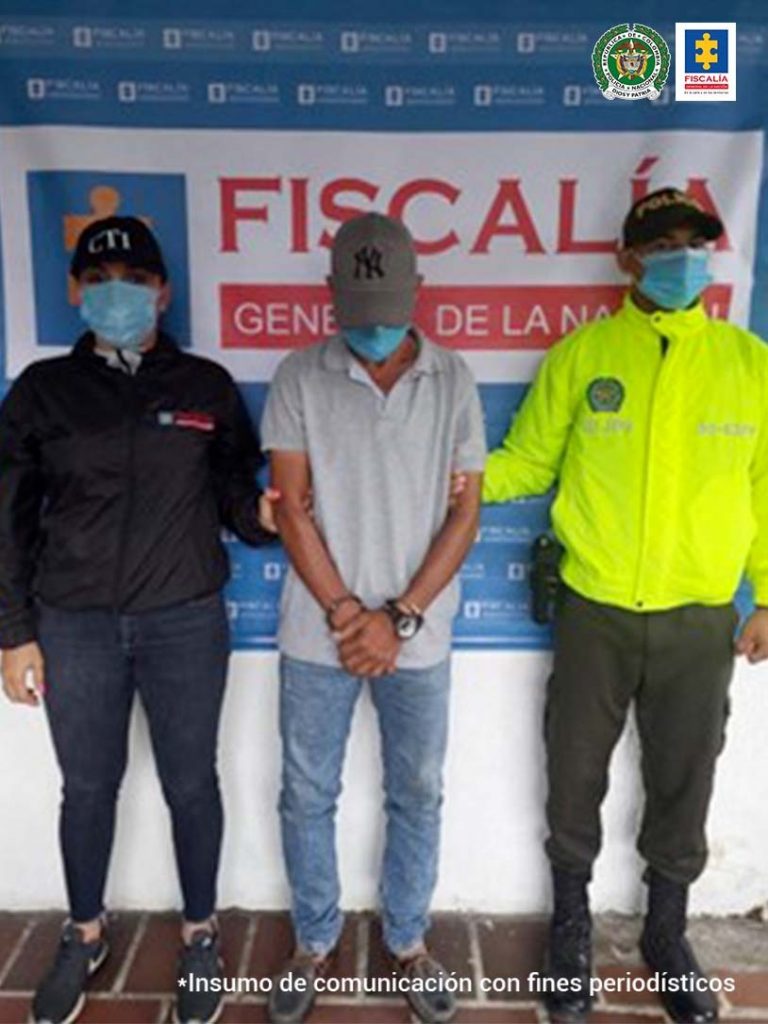 En la fotografía se aprecia el capturado junto a una investigadora del CTI y un uniformado de la Policía Nacional. En la parte posterior se observa el banner de la Fiscalía General de la Nación