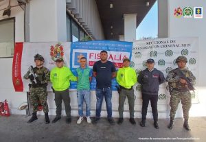 Se visualiza a los dos capturados junto a un investigador del CTI y uniformados del Ejército y Policía Nacional. Detrás de ellos los banners que identifican a la Fiscalía General de la Nación, Policía y Ejército Nacional.