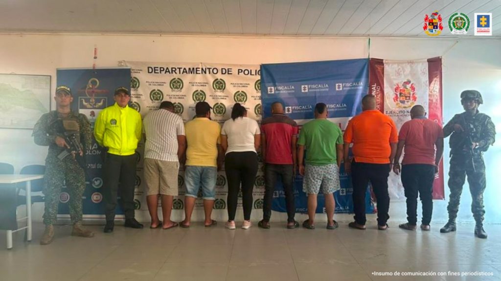 En la imagen se aprecia a los capturados de espaldas junto a uniformados de la Policía y Ejército Nacional. En la parte posterior se observa los banners que identifica a la Fiscalía General de la Nación y a la Policía y Ejército Nacional.