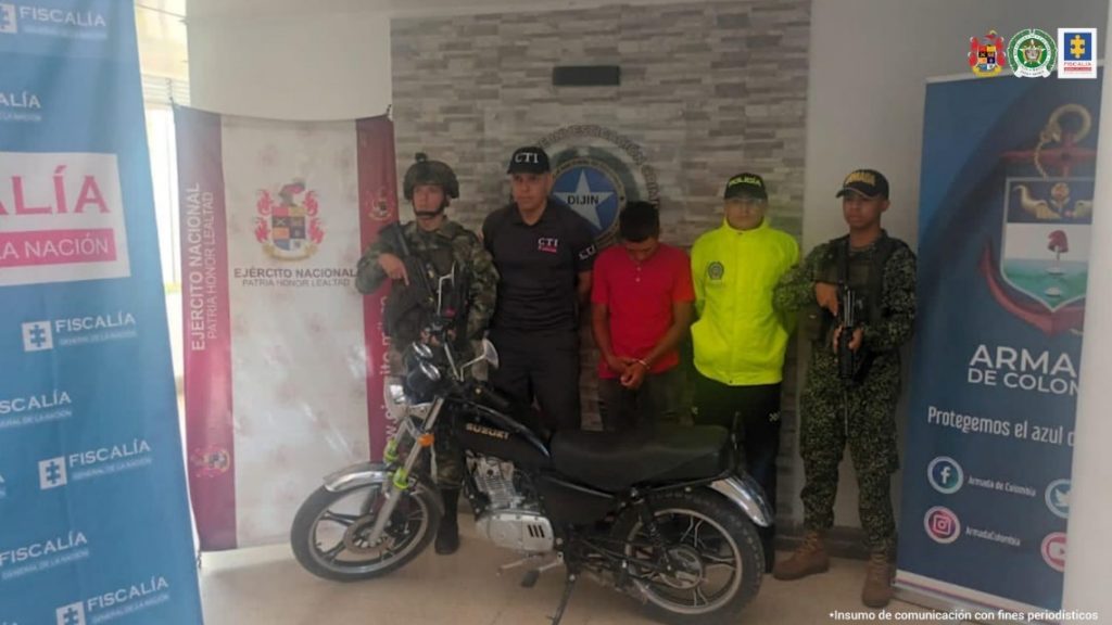 En la imagen se observa al procesado junto a un investigador del CTI y uniformados del Ejército, Policía y Armada Nacional. En la parte posterior se observa los banners que identifican a la Fiscalía General de la Nación, al Ejército, Policía y Armada Nacional.