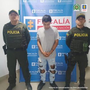 En la imagen se observa al capturado junto a dos uniformados de la Policía Nacional. En la parte posterior se aprecia el banner de la Fiscalía General de la Nación.
